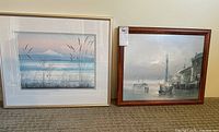 Photo showing two pieces of framed artwork: a coastal grass scene print in a light frame with matting on the left, and a boat dock scene painting in a brown wooden frame on the right.