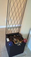 Photo of black resin garden pot with metal trellis stand, soil inside, and decorative garden accessories including wind chimes and two glass gazing balls.