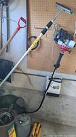 Wide view showing gasoline weed whacker with handle and fuel can, garden tools in large containers including shovel handles and watering cans, coiled hose on floor.