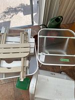 Two beige collapsible side tables, a white plastic side table with metal frame, a green watering can, a small green plant pot, and a brown ceramic plant pot placed indoors on tile floor.