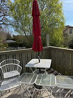 Full view of two white metal patio chairs, four rectangular glass-top side tables, square metal table with red umbrella, and umbrella stand on deck.