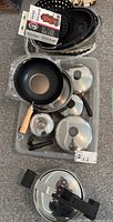 Overview of various cookware including frying pans, pots, percolator, and roasting pans in a plastic container and on the floor.