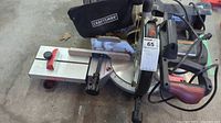Overall view of Craftsman mitre saw with base, angle adjustment gauge, and clamp holding wood piece, also showing circular saw and grinder behind