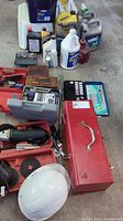 Overall lot showing toolboxes, electric angle grinder, white hard hat, assorted automotive fluids, various tool kits and socket sets arranged on floor.
