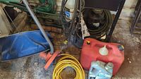 Photo showing vintage green gasoline lawn mower, orange string weed wacker, yellow extension cord, and red fuel cans on stained workshop floor