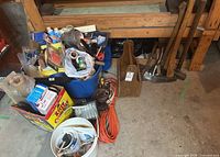 Photo shows a collection of various tools and supplies in buckets and boxes including painting tools, an orange worklight, and extension cords on concrete floor.