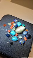 Mixed gemstone lot on black textured surface, showing various gemstone shapes and colors.