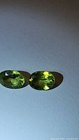 Two oval cut natural Peridot gemstones with apple green color shown side by side on a white background under direct light
