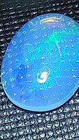 Close-up photo of natural Ethiopian opal oval cabochon showing blue and green flash within the stone on dark textured background.