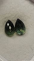 Close-up of two pear-shaped briolette cut sapphires in multiple shades of green and blue on a grey surface.