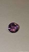 Close-up photo of single round cut fuchsia purple-pink fluorite gemstone on neutral background showing facets and transparency.