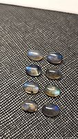 Eight oval cabochon natural labradorite gemstones arranged in two columns on a black textured surface, showing blue flashes of light.