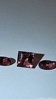 Three untreated natural pink tourmaline gemstones with high clarity, including one square cut and two oval cut stones, placed on a plain surface for detailed examination.