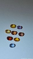 Eight oval cut sapphires of various rainbow colors displayed on flat surface under light showing clarity and color variety.
