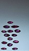 Top-down photo of 14 marquise cut rubies arranged against a light blue background showing their size and color variety in deep fuchsia red to pink-purple hues.