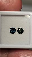 Two round cut sapphires in white cushioned display, showing blue-green coloration and good clarity for the stones.