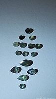 Photo showing a variety of sixteen natural alexandrite gemstones arranged on a flat surface. Various shapes and sizes, showing typical green to yellowish hues and some purple tint.