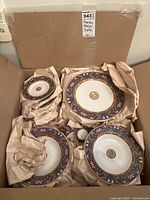 Box opened showing packed floral patterned porcelain dishes including plates and bowls with decorative multicolor rim.