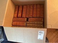 Photo showing a box containing approximately 16 antique leather-bound books with red-brown covers and visible wear.