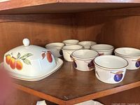 Covered butter dish with peaches on lid and plate, along with eight ramekins with fruit motifs in the background.