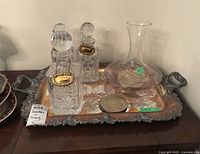 Shows the electroplated copper tray with ornate handles and three crystal decanters with metal tags labeled Scotch, Brandy, and Gin, plus an art glass style decanter.