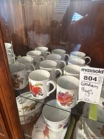 Photo showing a collection of approximately 12 Royal Worcester Evesham mugs in a display cabinet, each with distinct fruit illustrations and gold rims.