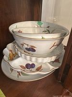 Stacked Royal Worcester Evesham serving dishes showing bowls, quiche pan, crudite platter, round serving plate, and oval platter with fruit motifs and gold trim.