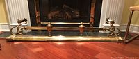 Full view of the brass fireplace fender laid out on wooden floor in front of a fireplace, showing overall length and design.
