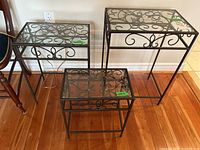 Front view of the three nesting tables showing the metal scrollwork and glass tops on a wooden floor.
