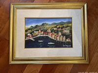 Full view of framed landscape painting labeled as Portofino, Italy by artist Mara Sanguineti, showing colorful coastal buildings, two sailboats, and green hills.