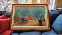 Full view of the framed oil painting showing two trees with blue-green foliage and a warm, colorful background, displayed on a couch.