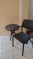 Photo showing black resin chair with perforated seat and backrest next to decorative black metal side table with ornate top and curved legs on tiled floor.