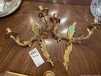 Photo showing both brass wall sconces resting on a wooden table, displaying their ornate leaf-shaped arms and two candle holders each.