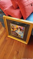 Framed colorful serialgraph artwork in a gold frame resting against cushions on hardwood floor.