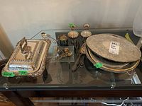 Photo of various silver plated trays, candlesticks, serving utensils, and glass plate arranged on a glass table.