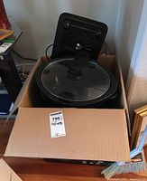 Box containing Crock Pot slow cooker with glass lid and black electric double hot plate seen behind in the box