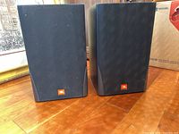 Front view of two JBL MR25 speakers with black cloth grille and JBL logo on a wooden floor.