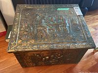 Front angled view of wood firewood box covered in embossed stamped metal scenes of people indoors playing cards or gambling along with other household items.