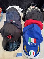 Photo showing 10 baseball caps stacked in two rows, varied colors including black, blue, red, and white, some with logos and embroidery such as Toronto Raptors and Italy flag.