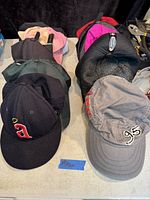 Top-down photo of 10 baseball caps arranged in rows showing various colors and designs including black cap with 'a' logo and gray cap with '95' embroidery