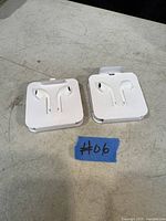 Photo showing two sets of Apple Lightning EarPods in their original white plastic holder packaging, placed on a surface with a blue tape marked #06.