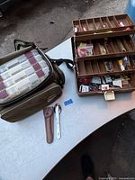 Two tackle boxes on a white table, one soft-sided green bag with plastic boxes visible inside, the other an opened metal box with trayed compartments filled with fishing lures and floats. Also, a fishing knife in sheath and a metal multi-tool opener set in front.