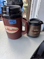 Two Tim Hortons thermos bottles displayed side by side. Large maroon thermos with handle on the left, smaller silver thermos with handle on the right.