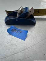 Blue metal small hand planer on a surface with measurement tape showing length, with blue tape labeled #54 beside it.