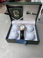 Open green presentation box displaying watch, golf balls, tee, and documentation.