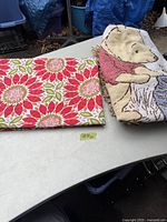 Photo of folded mid-century modern floral tablecloth and Winnie the Pooh blanket on a white table outdoors.