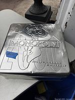 Square metal tin with embossed Monopoly logo and Mr. Monopoly character on lid, labeled Millennium Edition, small wear marks visible.