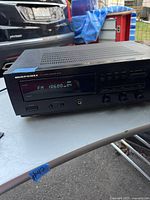 Front angled view of Marantz SR-48 stereo receiver on white surface with a car and blue tarp in background. Shows vent slots on top and front panel with display lit up.