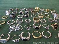 Multiple metal rings of various sizes and designs arranged on green cloth. Some rings feature black stones or enamel, twisted bands, gold and silver tones.