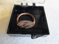 Ring displayed inside a black plastic square container, showing top view of engraved half oval top and corroded band